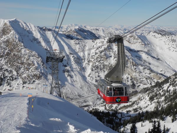 Snowbird's red aerial tram car suspended from its cables high above Little Cottonwood Canyon, skiers in helmets and goggles visible through the windows as it climbs toward the top station, with a steel tram tower standing on the ridge behind and snow-covered peaks, avalanche-scoured faces and dark stands of conifer falling away down the canyon.