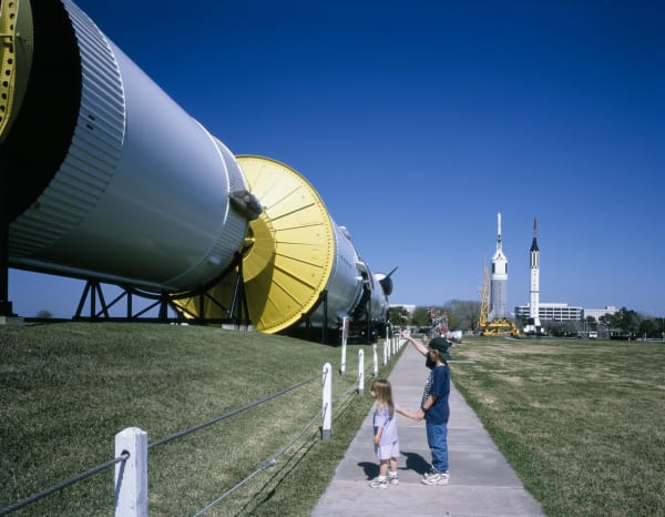 The stages of a Saturn V rocket lying end to end beside a walkway at Johnson Space Center, with two children looking on and smaller rockets in the distance.