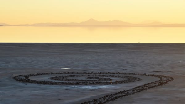 Robert Smithson's Spiral Jetty — a coil of black basalt rock curling counter-clockwise across pale, dry lakebed at the Great Salt Lake, with the water's edge lying well beyond the earthwork and low mountains on the horizon under a yellow sunset sky. Two small figures carrying buckets stand inside the innermost curve.