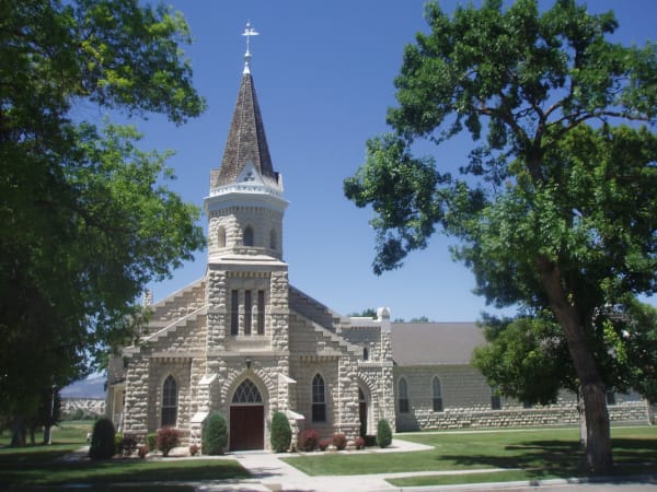 A church of pale rough faced limestone seen from across the street, a steep shingled spire and weathervane rising above an octagonal tower, gothic arched windows along the front, and a dark red door set under a pointed arch. Mature trees frame it on both sides and a clipped lawn runs to the sidewalk.