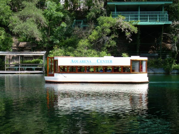A white glass-bottom boat on the clear green water of Spring Lake in San Marcos, with the old boat landing and trees behind.