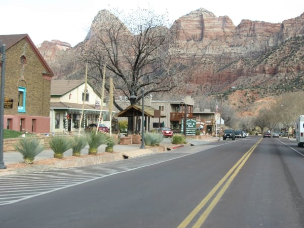 Zion Park Boulevard through Springdale in winter — a bare cottonwood over the sidewalk, shopfronts hung with garlands and signs for a gift shop and deli and the Pioneer Lodge along the right, parked cars and an RV further down the street, and the red and white cliffs at Zion's south entrance rising directly behind the town under overcast sky.