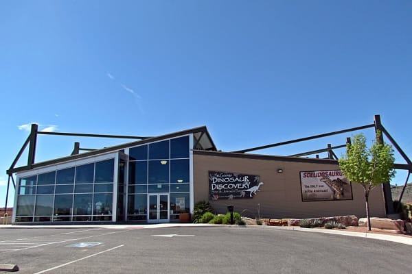 The Dinosaur Discovery Site museum at Johnson Farm — a low steel-framed hall with a tall glazed front and exposed roof struts, banners on the side wall reading ST. GEORGE DINOSAUR DISCOVERY SITE AT JOHNSON FARM and SCELIDOSAURUS, THE ONLY ONE IN THE AMERICAS, and an empty parking lot in the foreground under clear blue sky. The trackways themselves are inside the building and not visible.