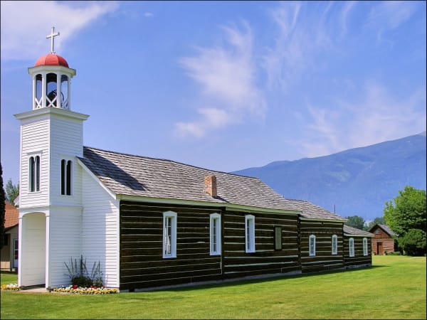 St. Mary's Mission chapel, dark squared logs under a shingle roof with a white clapboard bell tower topped by a cross, the Bitterroot Range beyond