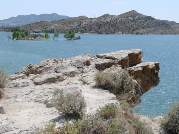 A pale rock shelf on the shore of Steinaker Reservoir, its lichen-stained blocks breaking away above blue-green water with sagebrush growing out of the cracks. A low wooded spit of cottonwoods stands out in the reservoir beyond, and dry juniper-dotted hills carrying transmission towers run along the far shore under a hazy white sky.