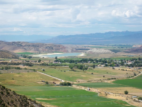 A wide green valley seen from high on one side: irrigated hayfields in strips of green and tan with sprinkler lines running, farmsteads and a small town spread among cottonwoods, and a pale turquoise reservoir set against white bluffs in the middle distance. Dry hills and a long blue range close the horizon.
