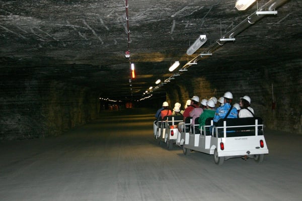 Visitors in hard hats ride an open tram down a long, lamp-lit tunnel cut through rock salt at Strataca.