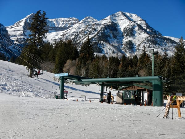 The base of a lift at Sundance — a green steel chairlift terminal under its canopy with a sign advertising complimentary guided tours, a small operator's hut with ski racks and poles beside it, a few skiers in dark clothing on the packed snow, and the snow-covered summit ridge of Mount Timpanogos rising behind the conifers under deep blue sky.