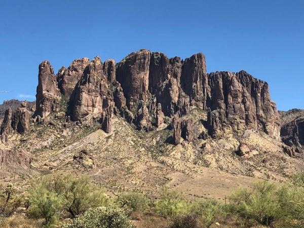 The west face of the Superstition Mountains — a long wall of columnar volcanic rock in warm brown and grey rising across the whole frame in buttresses and pinnacles, with pale talus chutes falling between them and scree gathering at their feet. A bajada of desert scrub, palo verde and cholla runs along the bottom of the frame beneath the wall, and the sky above is deep and cloudless.