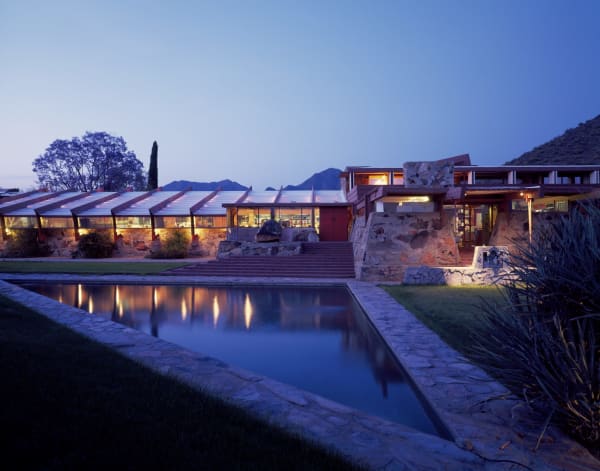 Taliesin West at dusk. Its low ranges are built of desert masonry — rough local stone set in concrete — under canted beams, with a long run of angled clerestory glazing lit warm from within and reflected in a rectangular pool that fills the foreground. Stone steps climb between the wings to a red door, flagstone terraces and strips of lawn edge the water, an agave stands at the right and a cypress and a bare tree at the left, and dark hills close the view under a deep blue twilight sky.