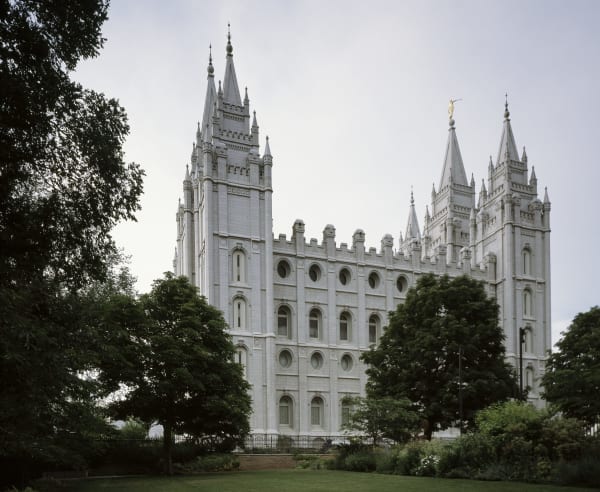 The Salt Lake Temple seen across a lawn from the south — grey granite walls with rows of oval and arched windows below a castellated parapet, six spires rising against an overcast sky with the gilded Angel Moroni on the tallest, and mature trees and a flowerbed in front of the iron perimeter fence.
