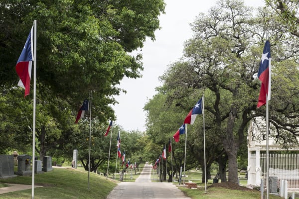 Texas flags on tall poles lining a long paved walk under live oaks at the Texas State Cemetery in Austin.