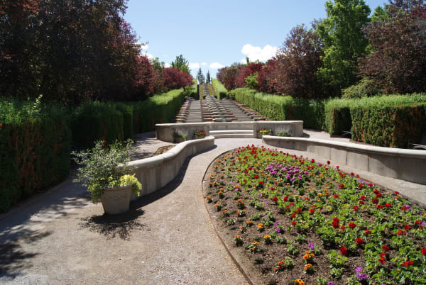 A formal garden at Thanksgiving Point — a curving gravel path between low rendered retaining walls, a crescent bed massed with red, orange and purple annuals in the foreground, clipped hedges and copper-leaved trees on both sides, and a long flight of stone steps with a water rill climbing the slope to the skyline beyond.