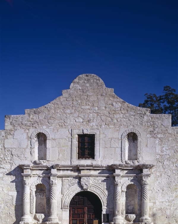 The weathered limestone facade of the Alamo church in San Antonio, with its carved doorway, empty niches and curved parapet against a deep blue sky.