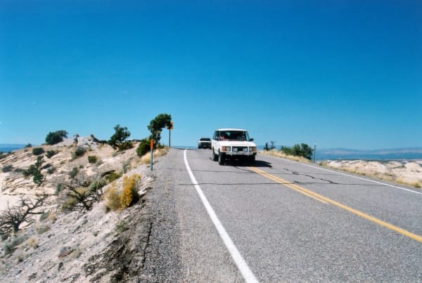 Highway 12 running along the spine of the Hogback in Utah — a white SUV oncoming across the center line, the ground falling away on both shoulders and junipers on the left.