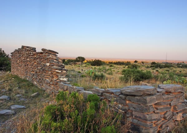 The low, crumbling limestone walls of the Pinery Butterfield stage station among grass and shrubs at dusk.