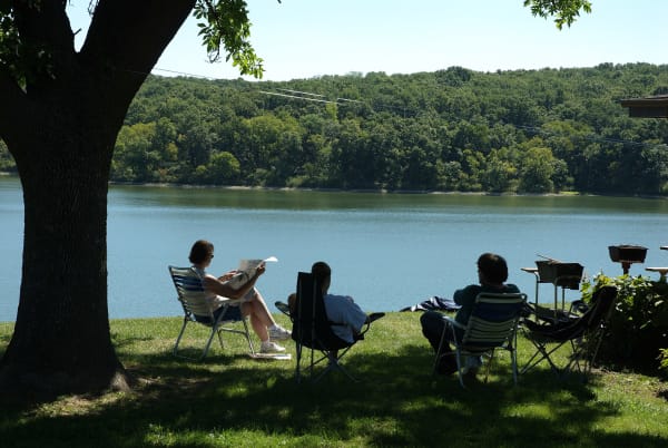 Visitors in lawn chairs under a shade tree on the shore of Forest Lake, with wooded hills across the water.