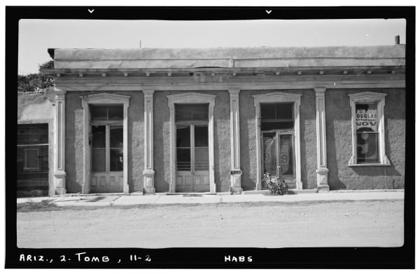 A Historic American Buildings Survey negative photographed in November 1937 and scanned with its black mount and index lettering in frame, reading ARIZ., 2. TOMB, 11-2 and HABS along the bottom edge. The plate is a window detail of the bank at Third and Allen Streets in Tombstone — four openings in a stuccoed front, three tall panelled doorways and a window at the right, moulded hoods over each, pilasters between them and a wooden cornice above, with the render fallen away in patches. A circus bill pasted in the right window advertises a Tuesday, November 9 date at Douglas. No street and no other part of the town is in the frame.