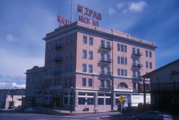 The Mizpah Hotel in Tonopah from the street corner, five storeys of pale brick above a rusticated stone ground floor, with iron balconies stepping down two faces, an arched entrance bay and a MIZPAH HOTEL sign in red letters carried on a steel frame above the roofline. A lower two-storey block with a cast-iron balcony adjoins it on the left, a pedestrian-crossing sign and parked cars stand at the kerb, and high cloud crosses the sky. The frame is scanned from a colour transparency and the whole image carries a strong blue cast.