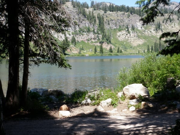 A small alpine lake ringed by conifers, seen from a shaded bank of pale boulders and packed earth in the foreground. A steep slope of grey limestone ledges and scattered spruce rises directly behind the far shore.