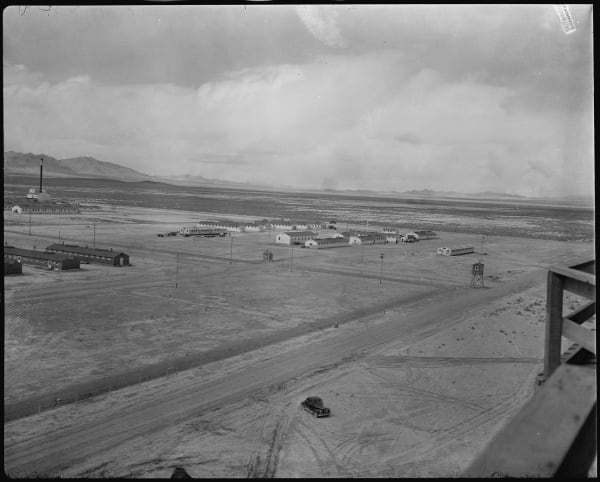 An archival War Relocation Authority photograph looking down over the camp from a raised vantage: long rows of low barracks and utility buildings standing on flat bare desert, unpaved roads, a fence line, and a watchtower raised on four braced legs alone on open ground. A single car sits on the road below and distant ranges close the horizon.
