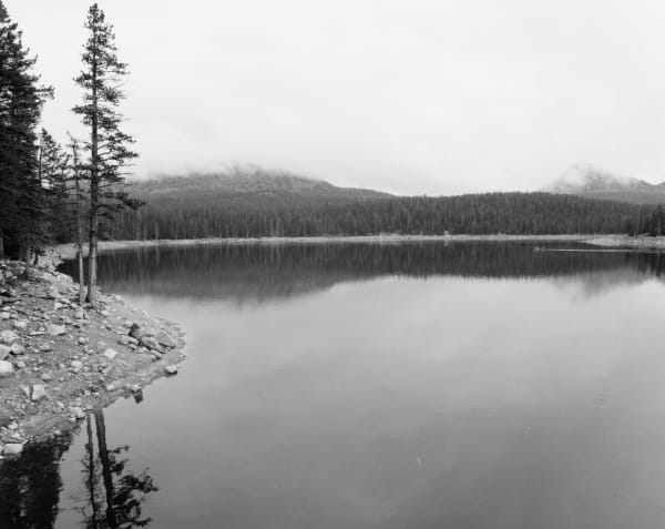 A black and white photograph of a still alpine lake, rocky shoreline and tall conifers in the left foreground, dense spruce forest along the far shore and cloud-covered ridges behind, the calm water holding a soft reflection under an overcast sky.