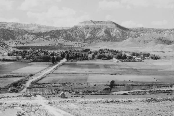 An archival black-and-white photograph of Tropic seen from the bench above — fenced hayfields laid out in a patchwork across the valley floor with a dirt road running down through them, a small board shed in the near field and cattle grazing beyond. The town's houses stand among cottonwoods and tall poplars, and the pale stepped escarpment of the plateau rim closes the view under cumulus.