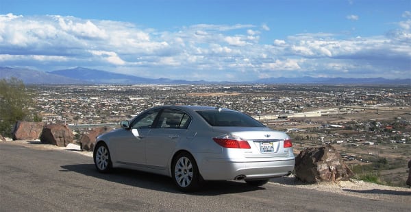 A silver sedan parked at a hillside pullout above Tucson, filling the centre of the frame in three-quarter rear view. Beyond it the city runs flat to the horizon — low houses and streets in the near ground, warehouse roofs and a rail yard in the middle distance, and a long range standing blue along the skyline under piled white cumulus. Broken red rock and gravel edge the pullout on either side. The photograph is composed on the car; the city sits behind it as backdrop, and nothing of the Old Pueblo or its farming ground along the Santa Cruz is legible at this distance.