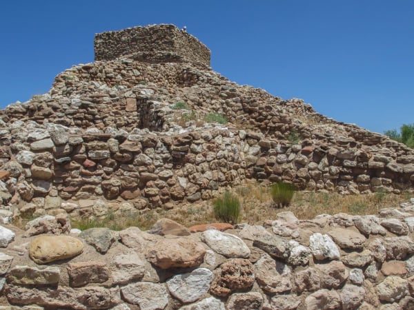 Tuzigoot from the foot of its hill, the pueblo rising in stepped courses of rough pale limestone and sandstone to a two-storey room block on the summit, wall lines running out along the slope on either side. Dry grass, rabbitbrush and bare stone fill the ground between them, a wall of large rounded cobbles crosses the immediate foreground, and a deep blue cloudless sky stands behind. The site was excavated and its walls raised again during the Depression, so part of what stands is reconstruction.