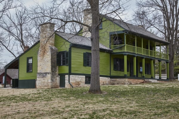 The green two-story White Haven house with its stone chimney and long porch under bare winter trees.