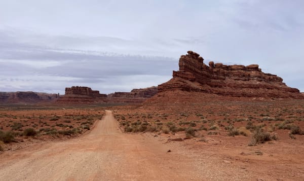 The graded dirt road through Valley of the Gods running away from the camera across red sand and saltbush, its wheel tracks worn pale through the rust-coloured surface. A broad red butte stands close on the right, banded in ledges and capped with a row of eroded knobs and blocks; further off, low mesas and a slender detached spire stand along the left horizon. The sky is flat grey overcast with a thin band of cloud lying across the tops of the buttes, and there is no vehicle and no person anywhere in the frame.