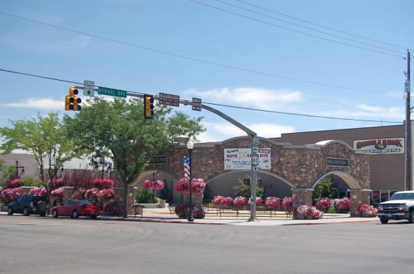 The corner of Vernal Avenue in Vernal, looking across at Cobble Rock Park — a river-cobble arcade of three round arches with COBBLE ROCK PARK lettered on panels at either end, hung with pink and white flower baskets and fronted by empty timber benches. A signal mast carries the street sign and brown arrows to Steinaker Reservoir, Red Fleet Reservoir and Flaming Gorge, a banner on the wall advertises ALIVE AFTER FIVE, and cars stand at the kerb under a hazy summer sky.