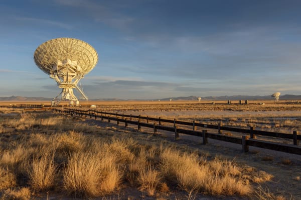 A white radio dish of the Very Large Array in low evening light, golden grass and a fence line in the foreground