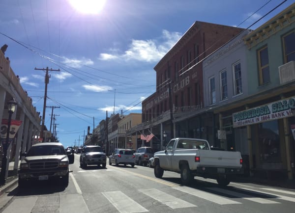 Traffic on C Street in Virginia City, pickup trucks and cars along a sunlit street of 19th-century commercial buildings with covered boardwalks, the brick Old Washoe Club of 1862 and a Ponderosa Saloon sign among the storefronts.