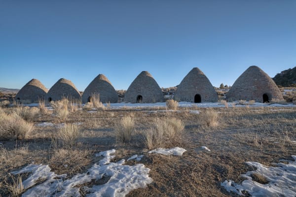 All six of the Ward Charcoal Ovens standing in a line — tall beehive cones of pale rough stone with arched doorways and small square vent holes, low winter sun raking across them from the right. Patches of snow lie between clumps of dry sagebrush in the foreground, and a low juniper-covered ridge closes the view behind.