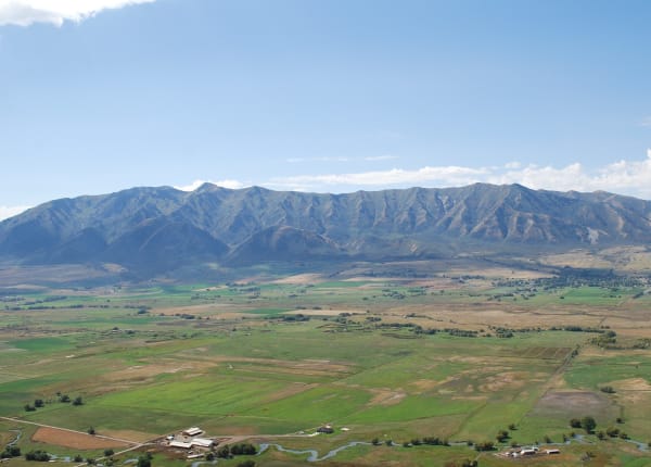 A long steep-sided mountain range rising abruptly from a flat green valley floor, its face cut into a row of parallel ridges and ravines. Irrigated fields, farm buildings and a meandering stream fill the foreground under a wide blue sky.
