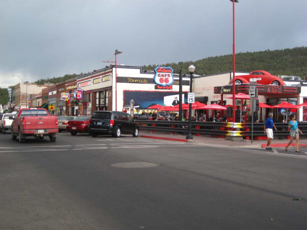 Route 66 through downtown Williams — a wide street with angled parking, a red pickup and a dark van at the kerb, and a row of low storefronts along the right under a wooded hill. Cruiser's Cafe 66 carries a large round Route 66 sign and a red car mounted above its entrance, with red patio umbrellas and a chrome-railed terrace in front, and neon signs run along the shopfronts beyond. Two people cross toward the camera at the right and diners sit out on the patio behind them.