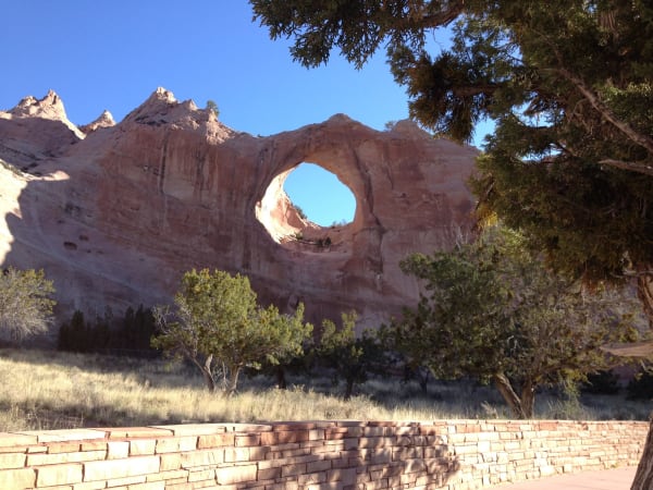 Tségháhoodzání, the arch that gives Window Rock its name — a near-circular hole worn clean through a fin of pale red sandstone, open sky showing through it, with the eroded ridge running away to jagged pinnacles at the left. Junipers stand across the middle ground and a mature juniper fills the upper right of the frame, dry grass covers the slope below, and a low wall of coursed sandstone slabs crosses the foreground. The arch is sacred to the Diné, one of the places where water is gathered for the Water Way, and no one climbs it.