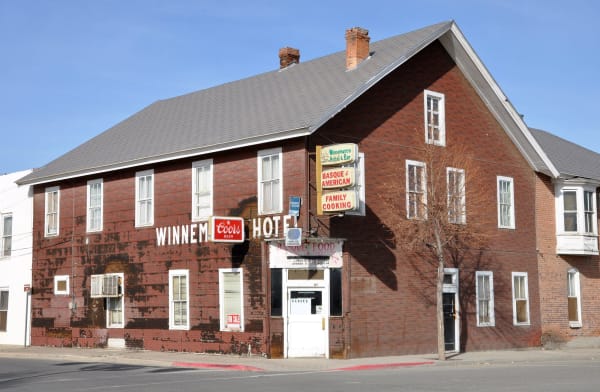 The Winnemucca Hotel, a two-storey red brick building with a steep grey roof and WINNEMUCCA HOTEL painted in white across the peeling brick with a Coors sign set between the words. A projecting panel sign reads Winnemucca Hotel & Bar, BASQUE & AMERICAN, FAMILY COOKING, a BASQUE FOOD awning covers the doorway below with the lunch and dinner hours lettered on it, and a CLOSED card and a FOR SALE notice show in the glass.