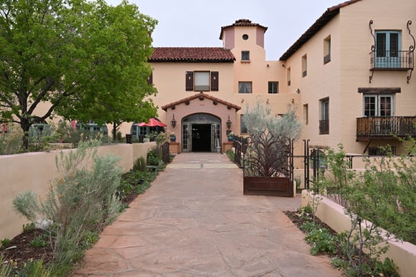 The garden approach to La Posada at Winslow, Mary Colter's 1930 hotel for the Santa Fe and the Fred Harvey Company — a flagstone walk running between low stucco walls and desert planting to an arched entrance under a red tile porch roof, with 1930 set into the plaster above the arch. The main block rises behind in pale stucco with a squat tower, shuttered casements, and wrought-iron balconies on the right-hand wing; cafe umbrellas stand at the left under a large tree. The sky is flat and overcast.