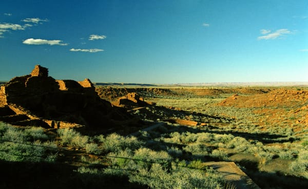 The masonry pueblo at Wupatki in low evening sun — red sandstone walls standing several courses high on a rise at the left of the frame, the room blocks stepping down the slope into shadow with a paved visitor path curving below them. Beyond, open grassland and dark red rock ledges run out to a far escarpment and a wide pale horizon under a deep blue sky. The building stone is the same red rock as the ground it stands on.