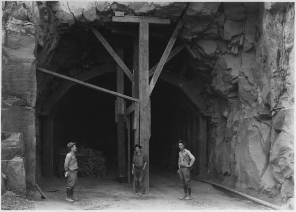 An archival black-and-white photograph of the Zion–Mount Carmel Tunnel under construction — the rough-blasted portal framed by heavy timber falsework and raking braces, stacked lagging timbers inside the dark opening, and three workmen in shirtsleeves, wide-brimmed hats and laced boots standing on the rubble apron in front of it. A hose runs out along the rock wall at right.