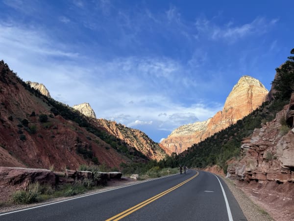 A park road in Zion at canyon level in low warm light — two lanes curving away with a double yellow line, red sandstone ledges along the right shoulder, and the canyon opening ahead between wooded slopes to sunlit orange cliffs and white-capped sandstone peaks under blue sky streaked with cirrus. Two cyclists ride on the shoulder in the middle distance.