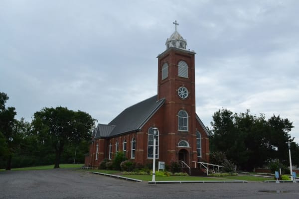 The red brick St. Joseph Catholic Church in Krebs, built in 1907, with its square bell tower under a grey sky.
