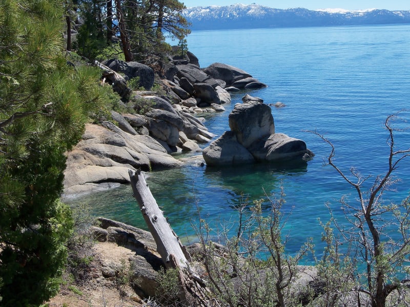 Large granite boulders in the clear turquoise shallows of Lake Tahoe's rocky Nevada east shore, with forested slopes and distant peaks under a blue sky.
