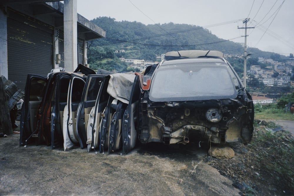 A stripped car and detached doors beside a hillside neighborhood