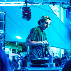 DJ playing under blue stage wash, moving heads and truss overhead at an outdoor show