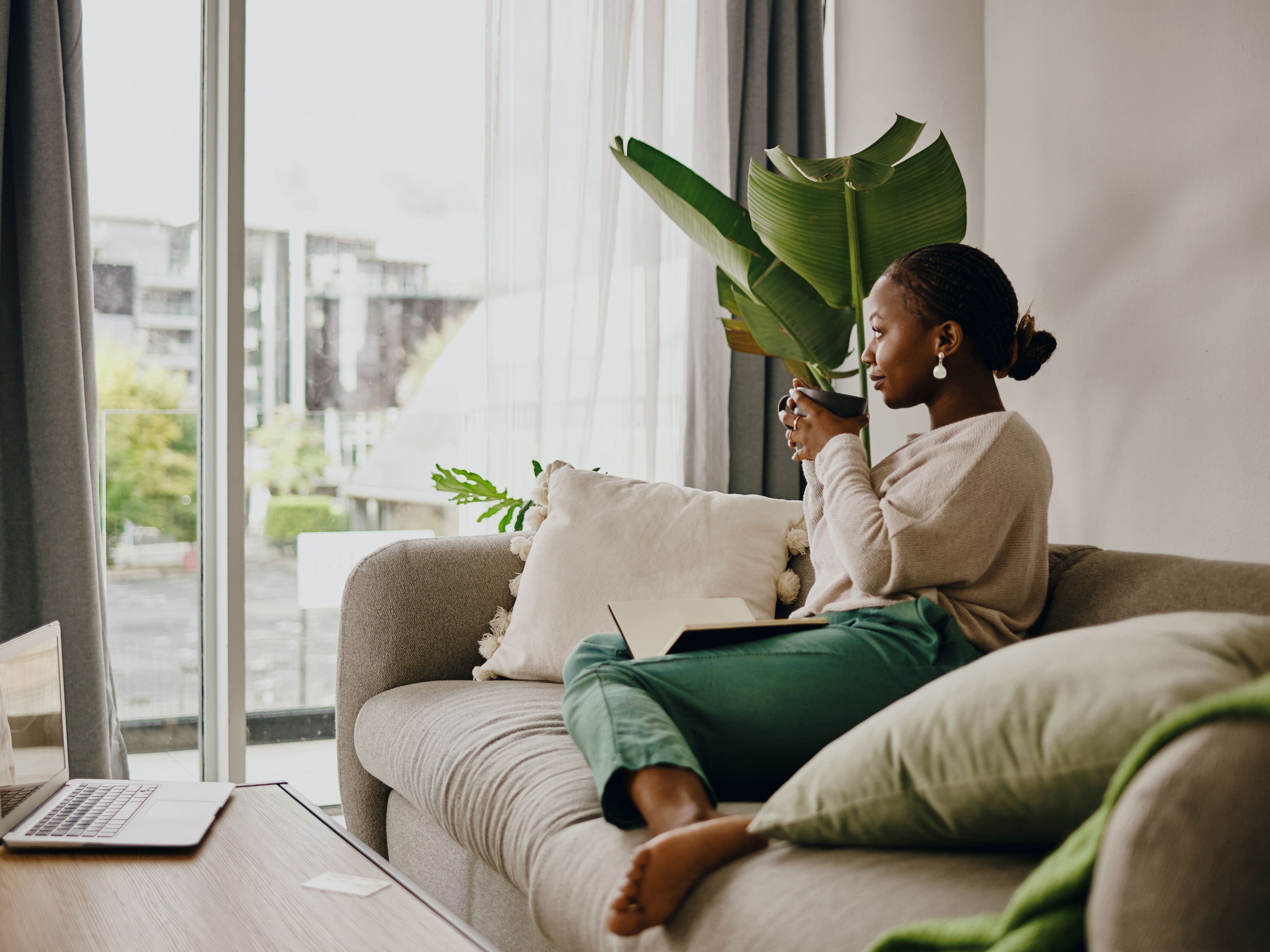 Woman sitting on her couch drinking tea next to a plant.
