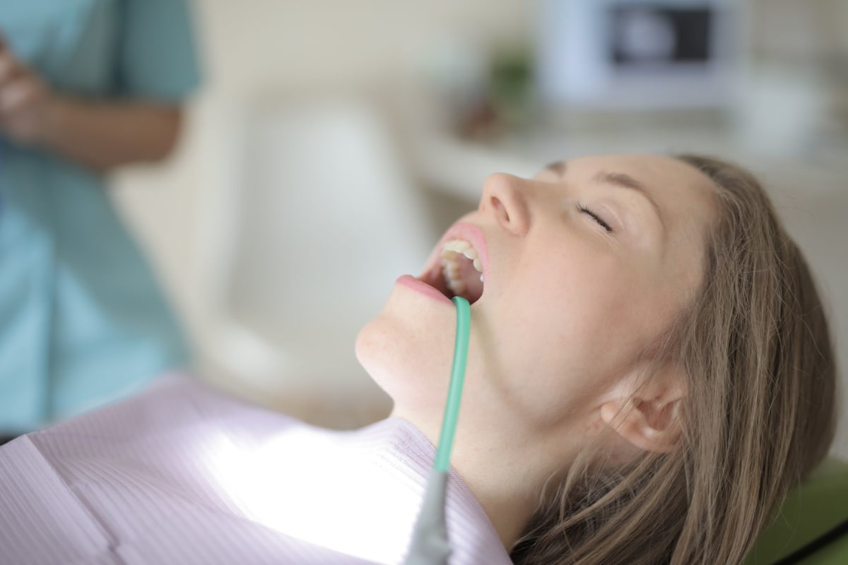 Side view of young female patient with saliva ejector in opened mouth sitting in dental chair while preparing for treatment in modern dental clinic