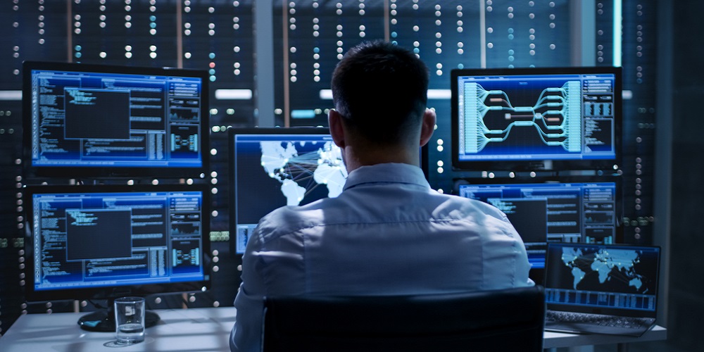 A man on a chair facing his back and looking at 6 screens with some lines of code and world maps